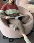 Person wearing brown slipper socks with the ankle area exposed, seated in a pink round chair reading a book.