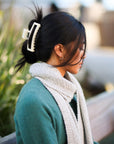 Woman with dark hair tied back with a white hair clip, wearing a green sweater and beige scarf, outdoors.
