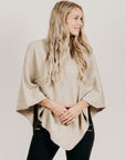 A woman wearing a beige Merinosilk poncho over a black top, paired with black pants. The poncho has a flowing design and she has long blonde hair.