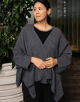 Woman wearing a gray poncho standing next to a plant indoors