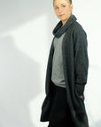 A woman standing, wearing a long gray cardigan with hidden side pockets over a gray sweater and black trousers.