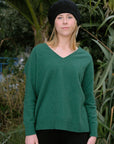 Person wearing a green sweater and black beanie standing outdoors with greenery in the background