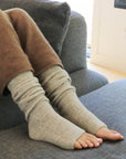 A pair of beige leg warmers worn on a person's legs, resting on a grey sofa.
