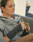 A person sitting on a sofa wearing a gray Merinosilk Tekapo wrist warmer while holding a cup.
