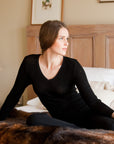 A woman sitting and wearing a long sleeve black merino thermal top with a V-neck.