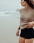 Woman wearing a beige sweater and black shorts standing on a beach.