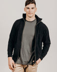 A man wearing a black Merinosilk pocket jacket with a full zip and standing with hands in pockets.