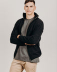 Man wearing a black zip jacket over a gray shirt with beige pants on a plain background