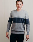 A man standing against a white background, wearing a gray and blue striped Merinosilk town sweater, paired with black pants.
