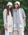 Two women wearing scarves with sheep and kiwi patterns standing in front of a green leafy background