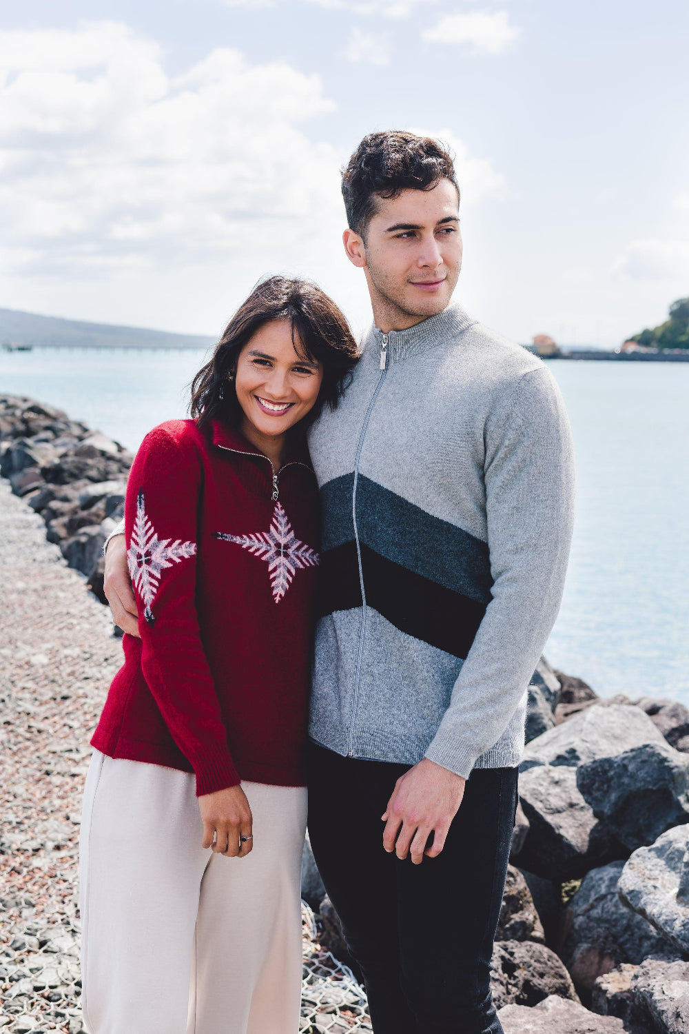 Two people standing on a rocky shoreline waring zip sweater with water in the background