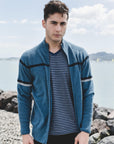 Man wearing a blue cardigan over a striped shirt by a body of water with rocks.