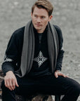 Man sitting on a log wearing a dark sweater with a patterned scarf and black pants.