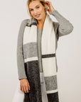 A woman wearing a gray and white striped alpaca scarf, paired with a gray sweater and white pants.