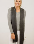 Woman wearing a long dark gray scarf over a light gray top and white pants on a plain background