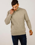 Man wearing a beige cable knit sweater and dark jeans on a plain background