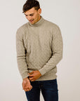 Man wearing a beige cable knit sweater and dark jeans against a plain background