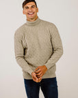 Man wearing a beige cable knit sweater and dark jeans against a plain background