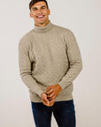 Man wearing a beige cable knit sweater and dark jeans against a plain background