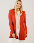Woman wearing a red cardigan over a beige top and white pants on a light background
