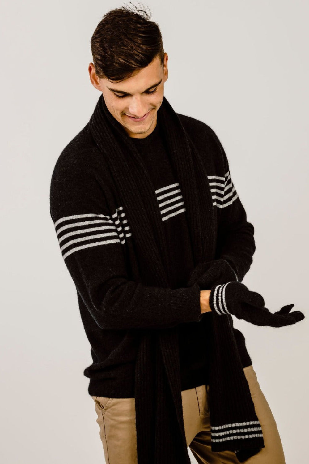 Man wearing a black sweater with white stripes and a scalf and matching gloves on a plain background