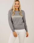 A woman standing, wearing a gray cashmere sweater with a crew neck and snow crystal design on the front, paired with white pants.