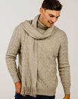 Man wearing a beige cable knit sweater and scarf against a plain background