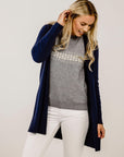 Woman wearing a navy cardigan, gray sweater, and white pants on a plain background
