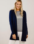 Woman wearing a navy cardigan over a gray sweater with white pants on a plain background