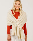 A woman wearing a cream-colored cashmere stole with a subtle texture, draped over her shoulders. She is also wearing a red top and white pants.