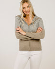 Woman wearing a gray zip-up hoodie and white pants against a plain background