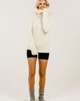 Woman wearing a cream cable knit sweater over a black pants with gray socks on a white background