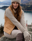 Woman in a beige vest and gloves sitting by a lake with mountains in the background