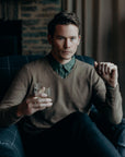 Man sitting in a chair holding a glass, wearing a brown sweater and dark jeans, with a blurred background.