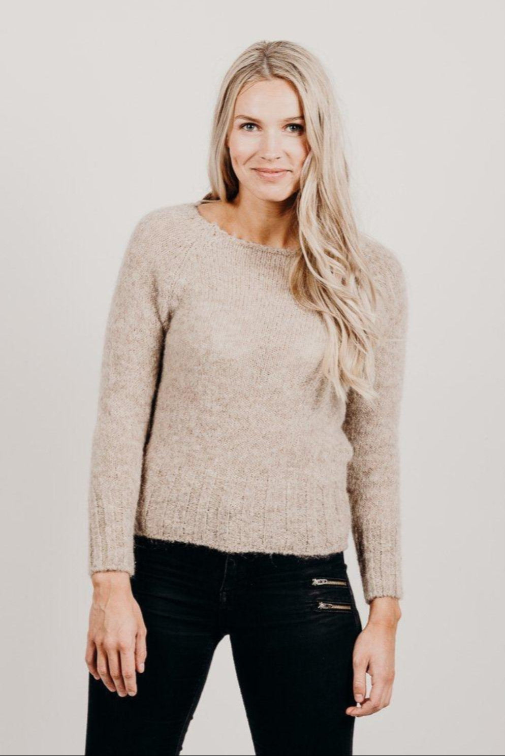 Woman wearing a beige sweater and black pants against a plain background