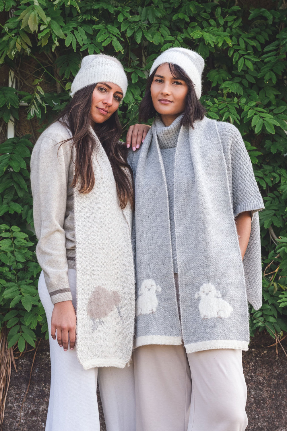 Two women wearing scarves with sheep and kiwi patterns standing in front of a green leafy background