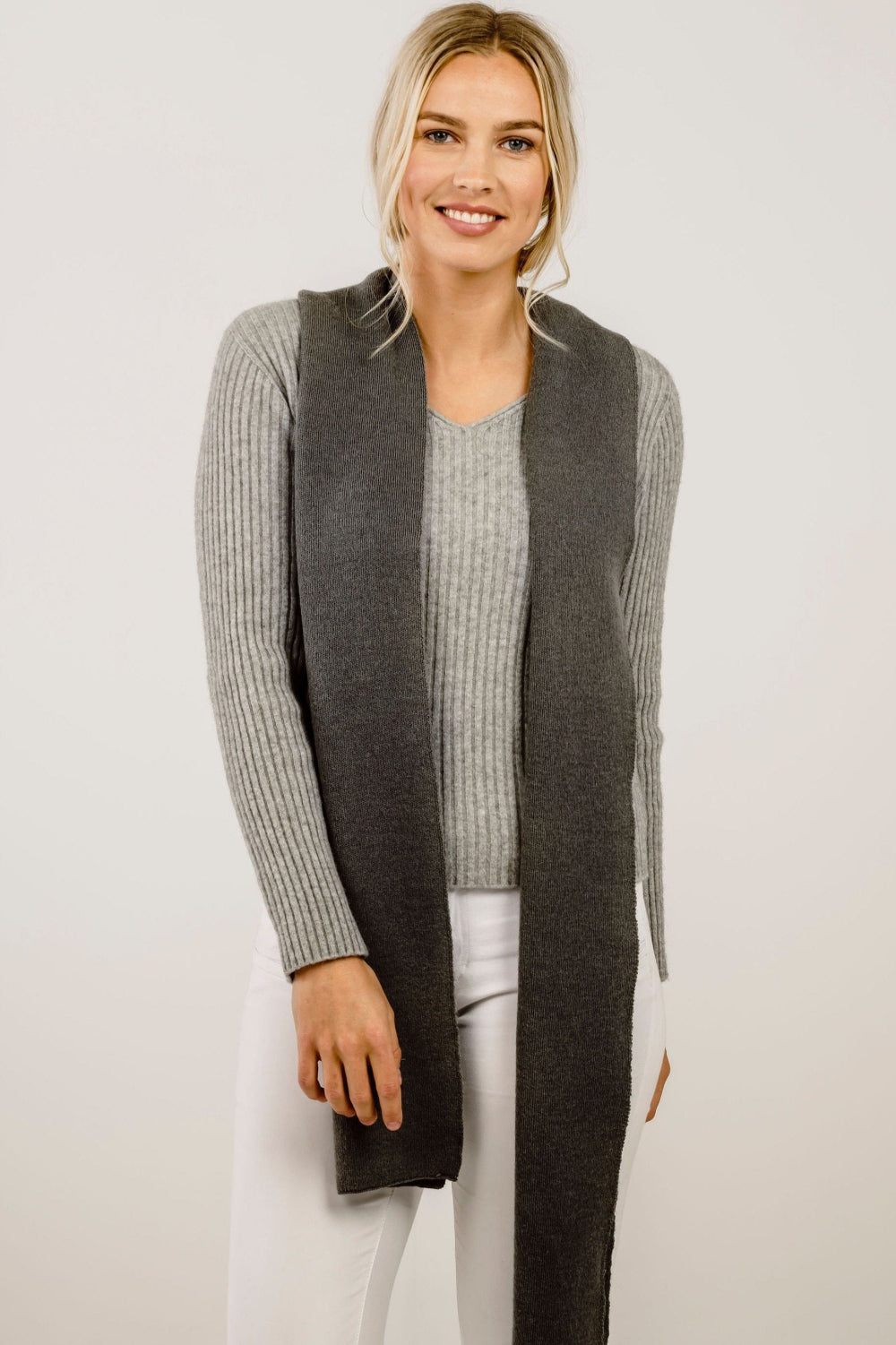Woman wearing a long dark gray scarf over a light gray top and white pants on a plain background